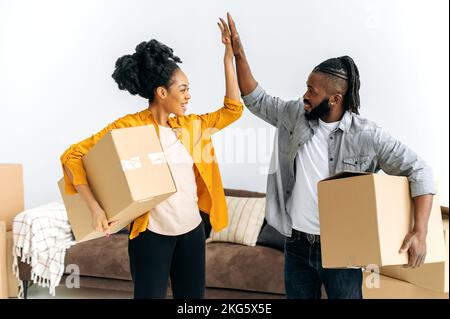 Gioioso sposato felice famiglia afro-americana coppia, tenendo scatole con le cose per la casa, stand in soggiorno, dare cinque a vicenda, sorridere, gioire nell'acquisto del proprio appartamento o casa Foto Stock