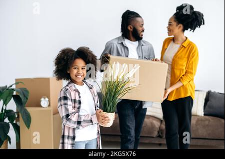 Felice piccola ragazza afroamericana di prescolare che tiene il flowerpot, genitori gioiosi che tiene le scatole di cartone, famiglia nera felice che si muove in nuova casa, comprando la loro prima sede. Concetto di rilocazione Foto Stock