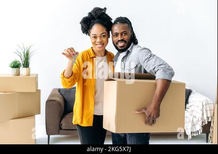 Una coppia amorevole sposata afro-americana si trasferisce nella sua nuova casa. Marito e moglie felici, in piedi nel loro nuovo salotto, uomo che tiene la scatola grande, donna che tiene le chiavi, che guardano la macchina fotografica, sorridendo Foto Stock