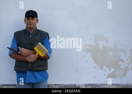 Sorridente handyman tenere pennello e vari strumenti che mostrano la differenza dopo il fissaggio di una casa parete da umidità e muffa. Foto Stock