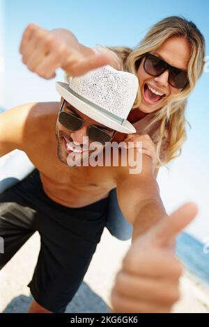 La vacanza migliore di sempre. una giovane coppia giocosa su una spiaggia soleggiata che dà i pollici fino alla macchina fotografica. Foto Stock