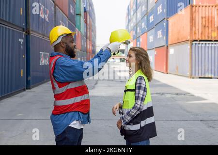 Un caposquadra con baffi e barba aiuta un apprendista femminile a indossare un elmetto. Entrambi indossano giubbotto di sicurezza riflettente. Ci sono contenitori nel w Foto Stock