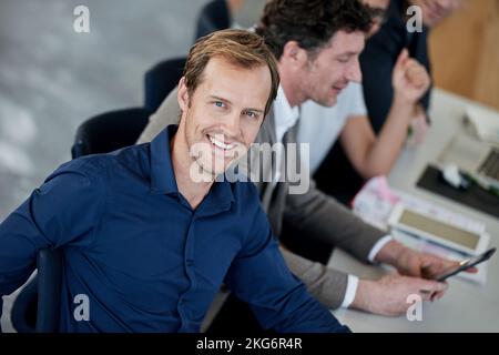 Non ho potuto chiedere un lavoro migliore. Ritratto di un bell'uomo seduto in ufficio con colleghi sullo sfondo. Foto Stock