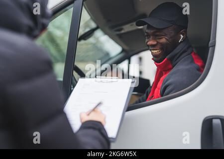 Concetto di pacchetti di posta e posta. Persona non riconoscibile in giacca nera che firma i documenti di consegna accanto al camion bianco. Bell'uomo afroamericano sorridente seduto sul sedile del conducente. Foto di alta qualità Foto Stock