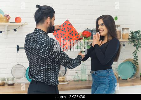 Un giovane amante inaspettatamente sorprende la sua bella ragazza con lunghi capelli castani scuri con un dono e una rosa rossa. Foto di alta qualità Foto Stock