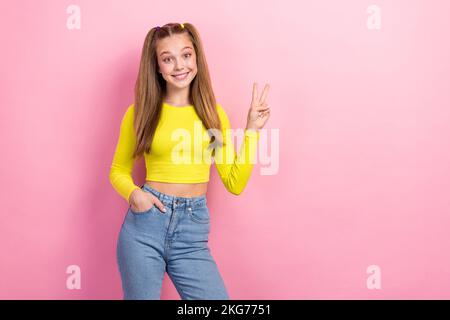 Foto di ragazza scolastica con coda di cavallo vestita giallo manica lunga che mostra segno v mano in tasca isolato su sfondo giallo Foto Stock