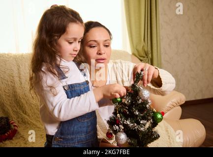 Madre amorevole e figlia amata che decorano insieme un albero di Natale, in attesa delle prossime vacanze invernali Foto Stock