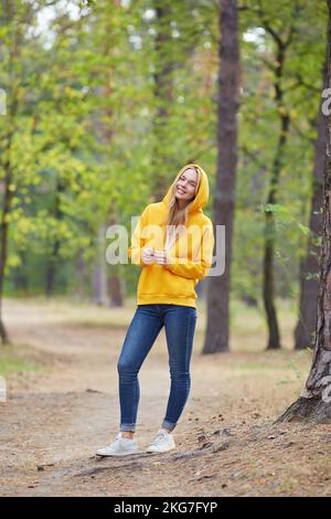 Bella giovane donna bionda in giallo felpa con cappuccio passeggiate nel parco. Attraente donna che passa il tempo attivo nel parco. Ritratto di una giovane donna gioiosa Foto Stock