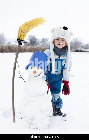 bambino allegro in tute calde, cappello a forma di orso polare si erge vicino al pupazzo di neve con secchio blu e scopa gialla. atmosfera invernale, rinculo stagionale all'aperto Foto Stock