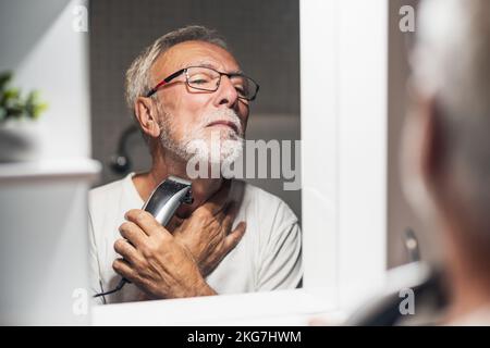 L'uomo anziano sta rifilando la barba con il rifinitore elettrico in bagno. Foto Stock