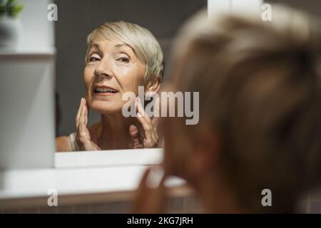 Donna matura che guarda il suo volto in bagno. Sta esaminando le rughe sulla sua pelle. Foto Stock