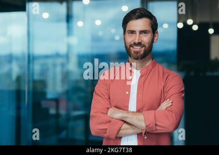 Ritratto di un felice e sorridente programmatore nel mezzo di un moderno ufficio, un uomo in occhiali e una camicia rossa sta guardando la macchina fotografica con le braccia incrociate. Foto Stock