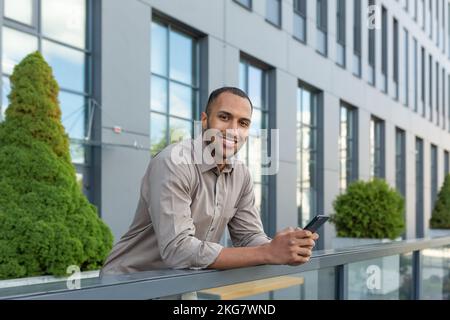 Un giovane bell'uomo d'affari latino-americano sta in piedi sulla veranda del centro ufficio, utilizzando un telefono cellulare, guardando la macchina fotografica, sorridendo. Foto Stock