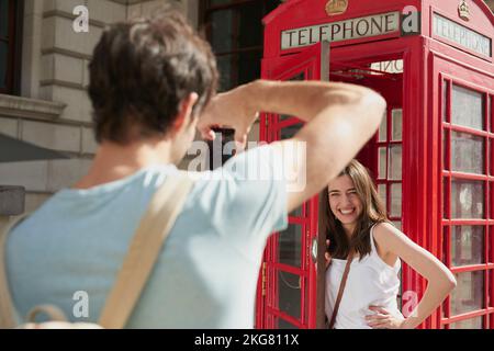 Cabina telefonica, smartphone e coppia in città per scattare foto in vacanza, in vacanza o in viaggio. Viaggi, fotografo e donna in chiosco rosso con l'uomo Foto Stock