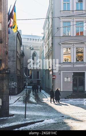 Riga, Lettonia - 20 2022 novembre: Coppia che cammina alla luce del sole nella strada del centro storico di riga, Lettonia, in inverno con bandiere, neve e ombre Foto Stock