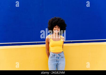 Bella giovane donna africana felice con capelli ricci afro passeggiando in città - allegro ritratto studente nero su sfondo colorato parete Foto Stock