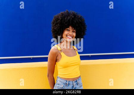 Bella giovane donna africana felice con capelli ricci afro passeggiando in città - allegro ritratto studente nero su sfondo colorato parete Foto Stock