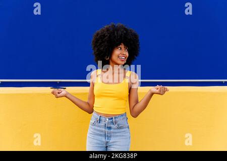 Bella giovane donna africana felice con capelli ricci afro passeggiando in città - allegro ritratto studente nero su sfondo colorato parete Foto Stock