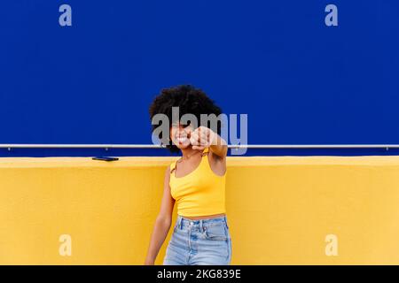 Bella giovane donna africana felice con capelli ricci afro passeggiando in città - allegro ritratto studente nero su sfondo colorato parete Foto Stock