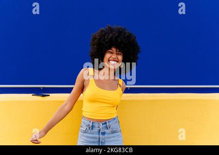 Bella giovane donna africana felice con capelli ricci afro passeggiando in città - allegro ritratto studente nero su sfondo colorato parete Foto Stock