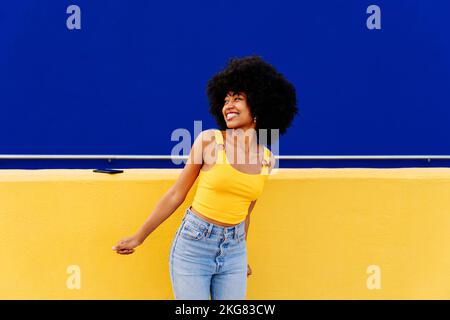 Bella giovane donna africana felice con capelli ricci afro passeggiando in città - allegro ritratto studente nero su sfondo colorato parete Foto Stock