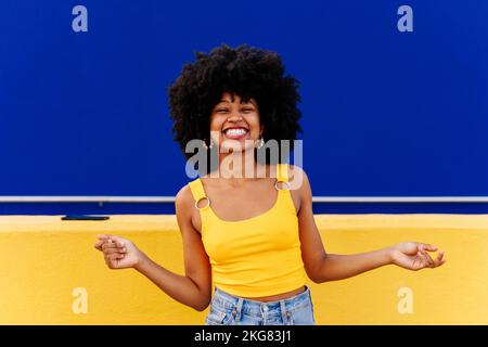 Bella giovane donna africana felice con capelli ricci afro passeggiando in città - allegro ritratto studente nero su sfondo colorato parete Foto Stock