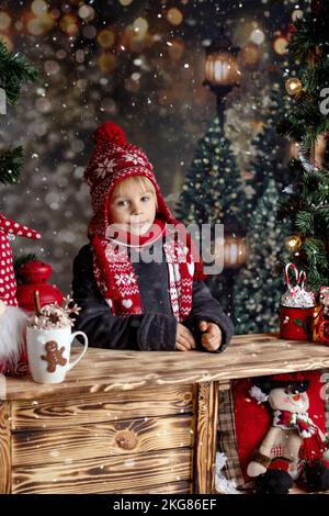Carino bambino, biondo ragazzo, in piedi nella neve su un piedistallo di legno, decorato per Natale, bere cioccolata calda e mangiare biscotti Foto Stock