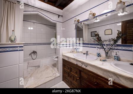 Bagno in stile vintage con ripiani in marmo, lavelli doppi, un grande specchio con sfumature e una vasca tra piastrelle bianche Foto Stock