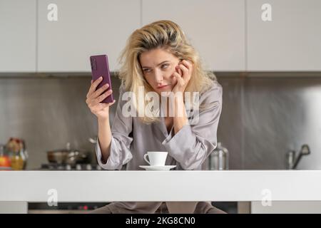 Ragazza sconvolta con il telefono bere vino in cucina. Mattina di bella donna. Signora triste che usa lo smartphone. Concetto di social media. Foto Stock