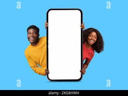 Sorridente coppia di millennial afro-americani che si scoppia da un enorme smartphone con schermo vuoto, consiglia blog Foto Stock