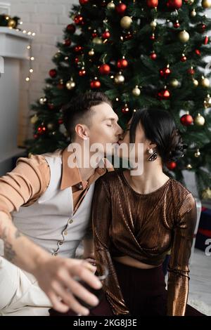 Uomo e donna asiatica con gli occhi chiusi baciandosi mentre si siede sotto l'albero di Natale Foto Stock