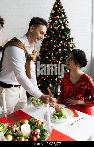 Donna asiatica incinta che guarda marito felice che serve insalata di verdure vicino candele brucianti a cena romantica di Natale Foto Stock