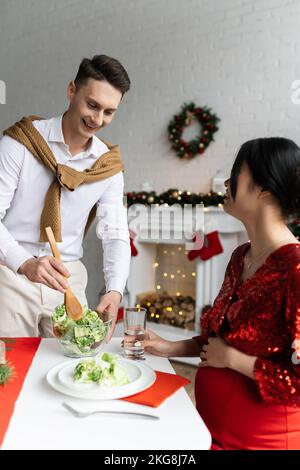 donna asiatica incinta seduta con un bicchiere d'acqua vicino al marito sorridente tenendo una ciotola con insalata di verdure Foto Stock