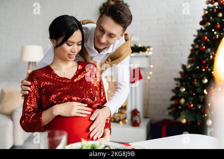 Uomo sorridente che tocca il pancino di una donna asiatica incinta mentre festeggia il Natale a casa Foto Stock