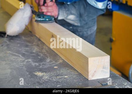 L'operaio di legno professionale rettifica la barra di legno sul banco di lavoro in un'officina di Falegnameria con l'attrezzo. Il lavoratore di mezza età in tute lavora legno con smerigliatrice elettrica. Flusso di lavoro reale... Foto Stock