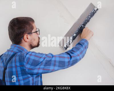 Mano di un giovane uomo adulto che usa spatola e parete bianca intonacata con stucco di finitura fresco. Primo piano. Lavori di riparazione di casa. Foto Stock