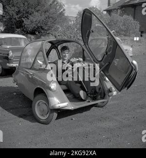 1960s, storico, fuori in un parcheggio di ghiaia, un uomo seduto all'interno di una piccola auto dell'epoca, una bolla auto, una BMW Isetta, che aveva una porta di apertura anteriore, Wycombe, Inghilterra, Regno Unito. Costruita per la prima volta da ISO Spa in Italia nel 1953, questa piccola auto è stata costruita su licenza in diversi paesi, tra cui BMW di Germania fino al 1962 e nota come la 'Bubble Car'. Foto Stock