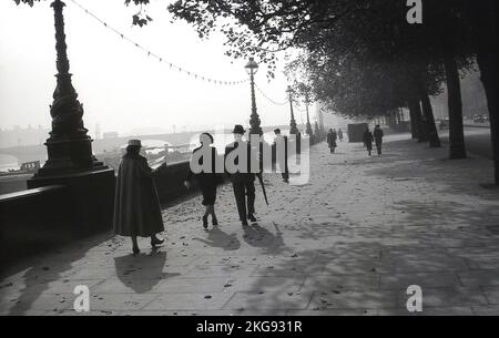 1950s, storico, un Signore e una signora elegantemente vestiti, entrambi indossati ha, camminando lungo l'ampio marciapiede a Victoria Embankment, una passeggiata lungo il fiume Tamigi, Westminster, Londra, Inghilterra UK. Foto Stock