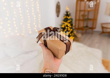 La mano femminile sta tenendo un regalo decorato, albero di Natale sfocato, presente avvolto, celebrando e dando concept.Christmas zero rifiuti. Confezione regalo Foto Stock