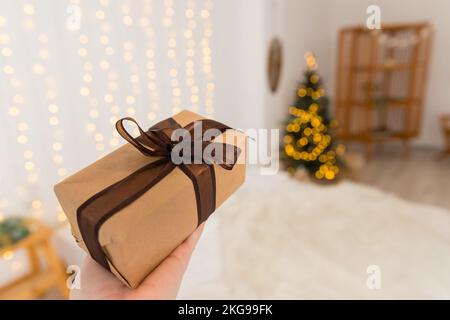 La mano femminile sta tenendo un regalo decorato, albero di Natale sfocato, presente avvolto, celebrando e dando concept.Christmas zero rifiuti. Confezione regalo Foto Stock