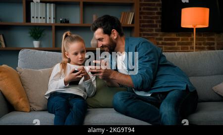 L'uomo caucasico e la bambina del capretto si siedono insieme nel paese tengono il telefono moderno che naviga il soddisfare di mezzi gioca i giochi in linea adulto dad insegnano l'uso della figlia del bambino Foto Stock