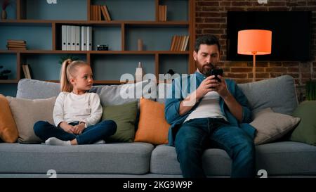 Annoiato bambino ragazza che riposa a casa con padre papà giovane utilizzando il telefono cellulare piccolo capretto bisogno di attenzione guardando papà prendere moderno gadget giovane famiglia Foto Stock