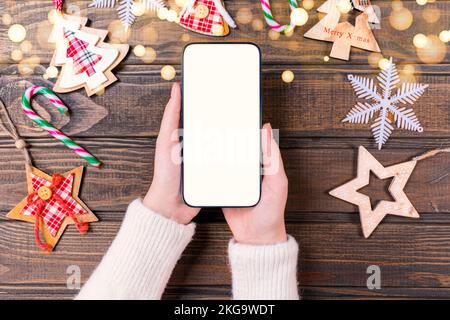 Primo piano donna che tiene il telefono con schermo bianco vuoto mockup vicino decorato albero di festa, festeggiamento di Natale, shopping cliente online, acquisto g Foto Stock