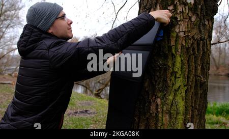 Uomo appendere pannello solare su albero per caricare smartphone da cavo multiuso, power bank, orologi, computer portatili sfondo natura. Energia pulita per l'uso in Foto Stock