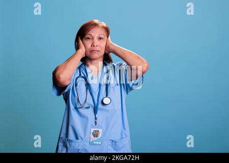 Infermiera asiatica tranquilla con stetoscopio che copre le orecchie con espressione seria durante la visita di controllo. Assistente medico che fa tre scimmie saggio simbolo. Non sentire il male concetto. Foto Stock