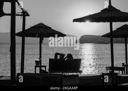 IOS, Grecia - 13 settembre 2022 : Vista di una coppia sdraiata sui lettini, bevendo vino e godendosi il tramonto arancione stupefacente in Grecia di iOS Foto Stock