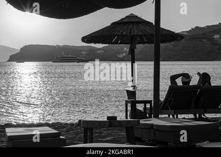 IOS, Grecia - 13 settembre 2022 : Vista di una coppia sdraiata sui lettini, bevendo vino e godendosi il tramonto arancione stupefacente in Grecia di iOS Foto Stock