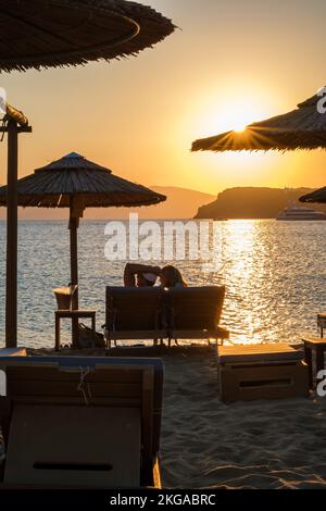 IOS, Grecia - 13 settembre 2022 : Vista di una coppia sdraiata sui lettini, bevendo vino e godendosi il tramonto arancione stupefacente in Grecia di iOS Foto Stock