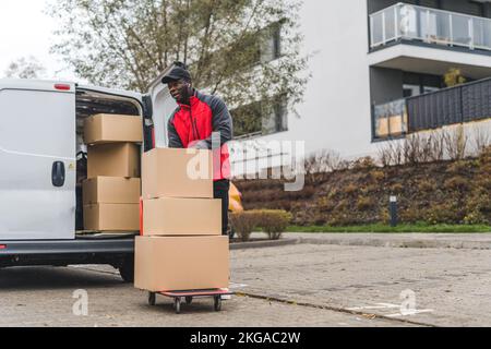 Foto completa all'aperto di un addetto alla consegna afro-americano che utilizza un camion manuale per consegnare scatole di cartone. Furgone di consegna bianco con tronco aperto pieno di pacchi. Edificio moderno sullo sfondo. Stagione autunnale. Foto di alta qualità Foto Stock