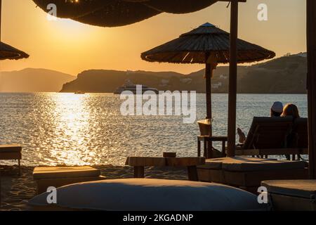 IOS, Grecia - 13 settembre 2022 : Vista di una coppia sdraiata sui lettini, bevendo vino e godendosi il tramonto arancione stupefacente in Grecia di iOS Foto Stock